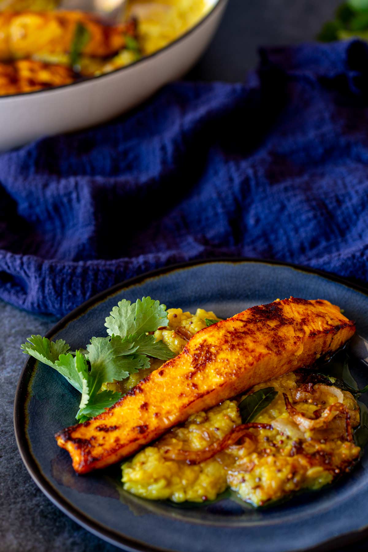 A close-up of a plate featuring a seared salmon fillet served on a bed of lentil daal, with fresh cilantro leaves on the side. The background shows the serving dish with more salmon daal.