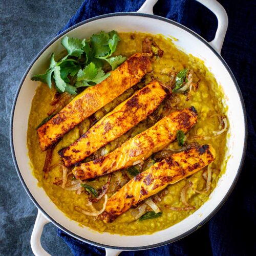 Seared salmon on top of golden lentil daal in a white skillet on a blue table