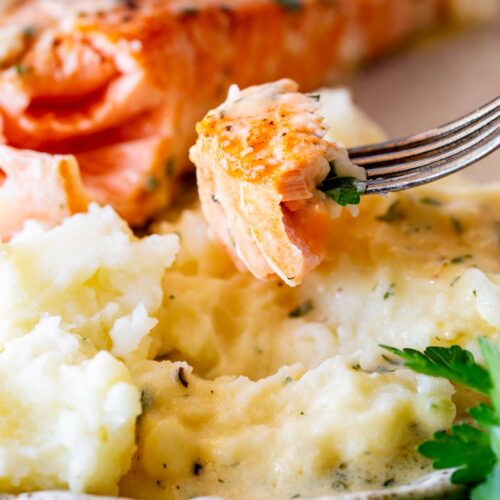 A close view of salmon with creamy herb sauce, partially flaked on a fork, next to mashed potatoes. A touch of fresh parsley adds color.