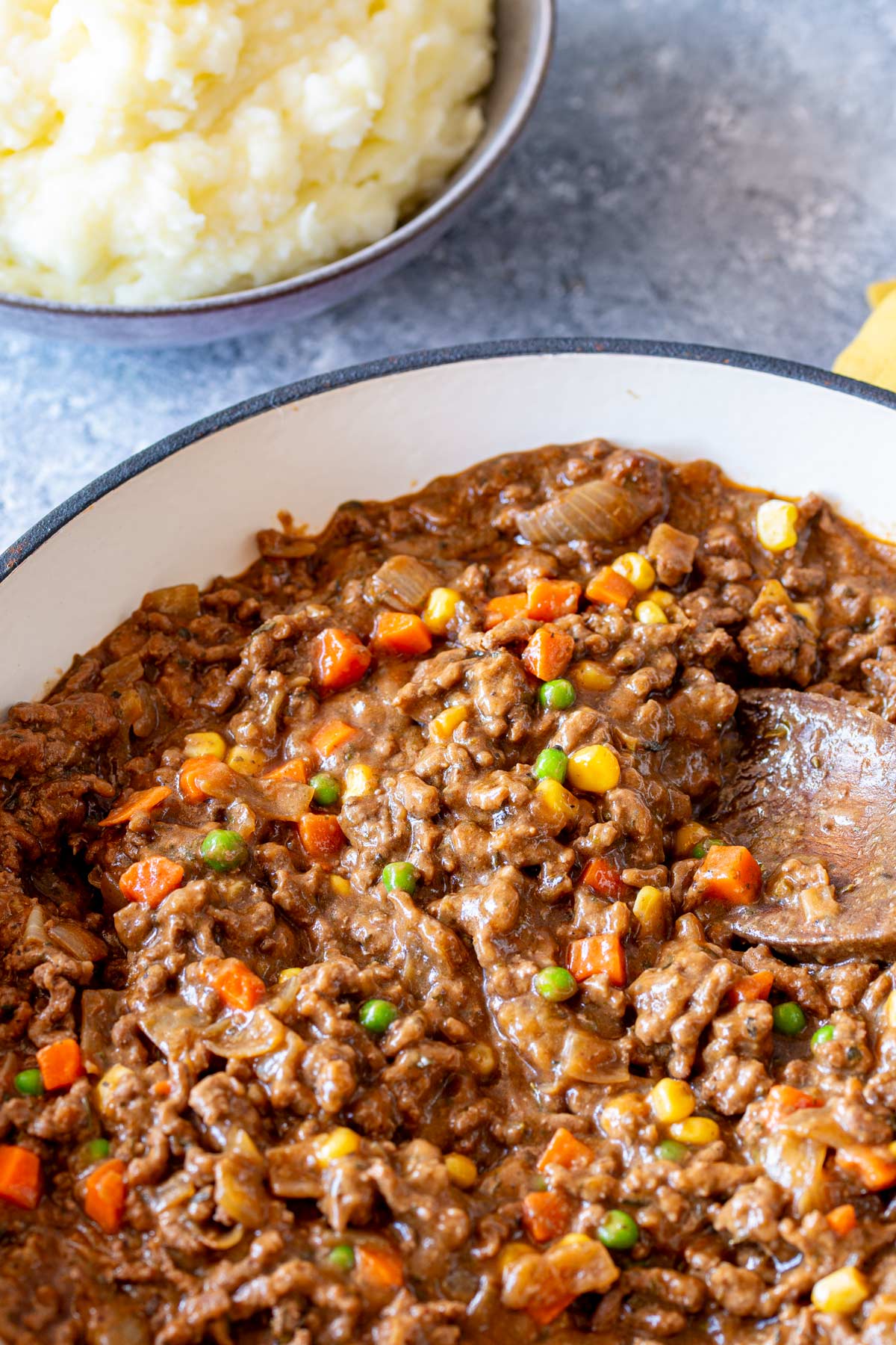 A large skillet filled with gravy mince cooked with carrots, peas, and corn, served with mashed potatoes.