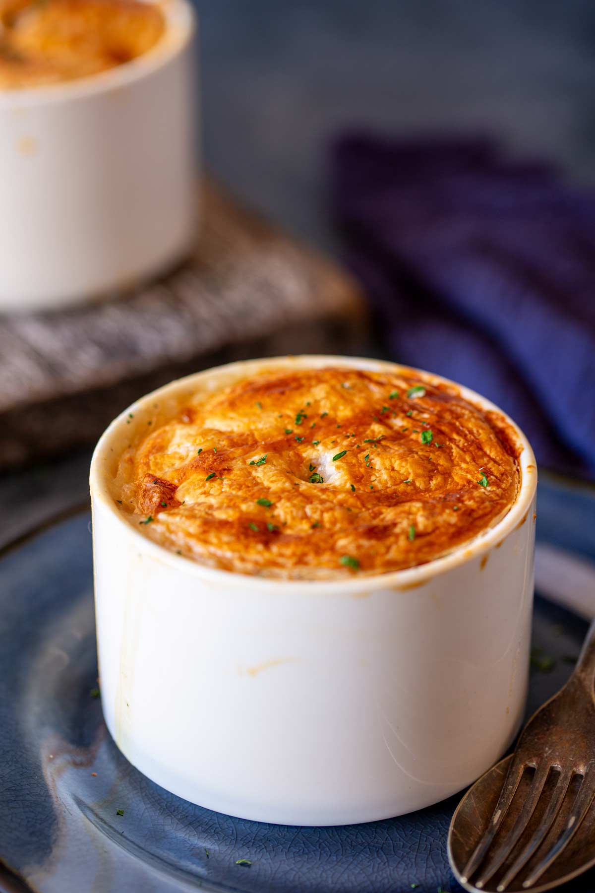 a white ramekin with a party top, sprinkled with parsley and sat on a blue plate with a rustic spoon and fork