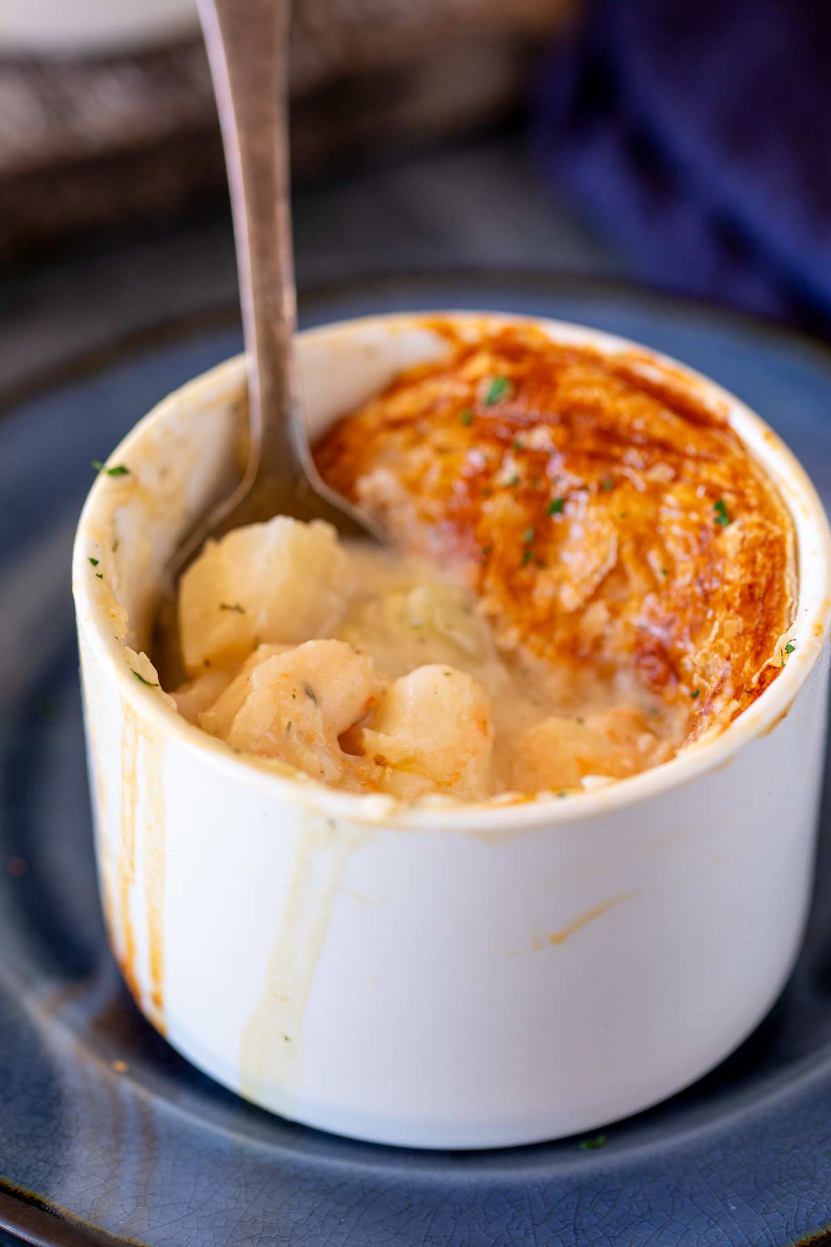 A spoon dipping into a seafood pot pie, revealing chunks of seafood and a rich, creamy sauce under a golden pastry top, served in a white ramekin.
