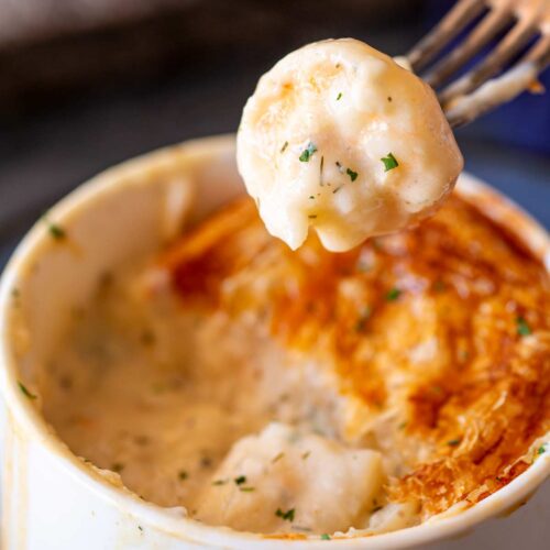 A forkful of creamy seafood filling with hints of parsley lifted from a ramekin, showcasing the flaky top layer of a freshly baked pot pie