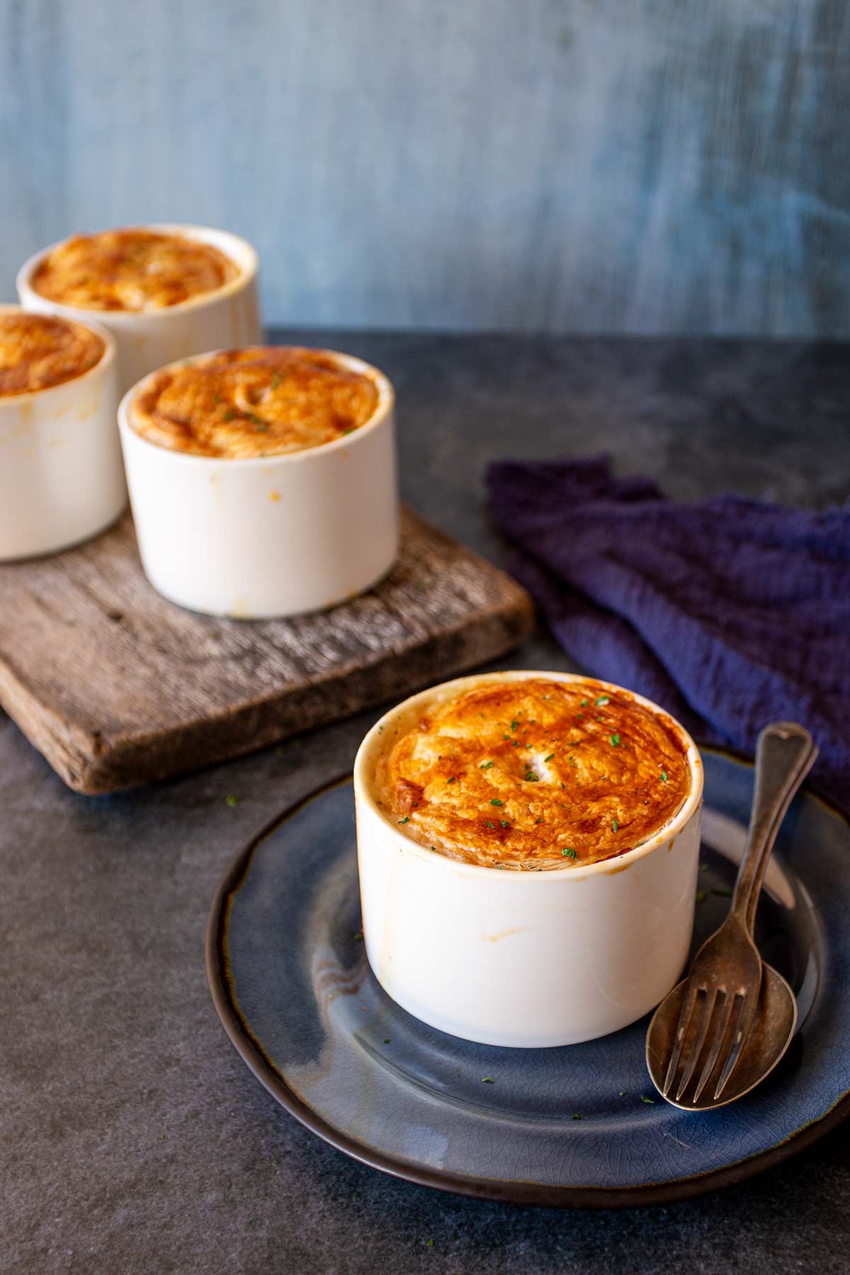 Four individual seafood pot pies in white ramekins with golden-brown, flaky pastry tops, three are on a rustic wooden board one is on a blue plate.