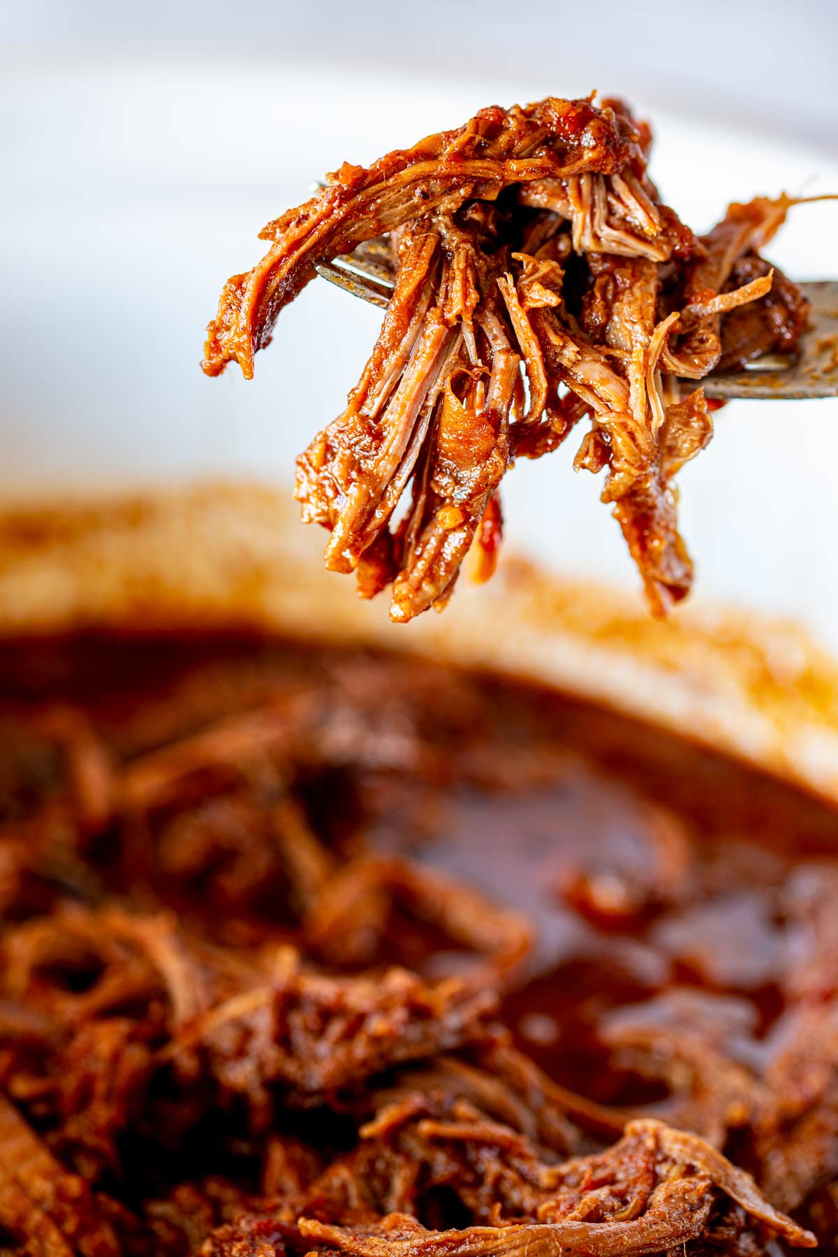 Shredded strands of BBQ beef held up by a fork over a bowl of saucy, slow-cooked beef.