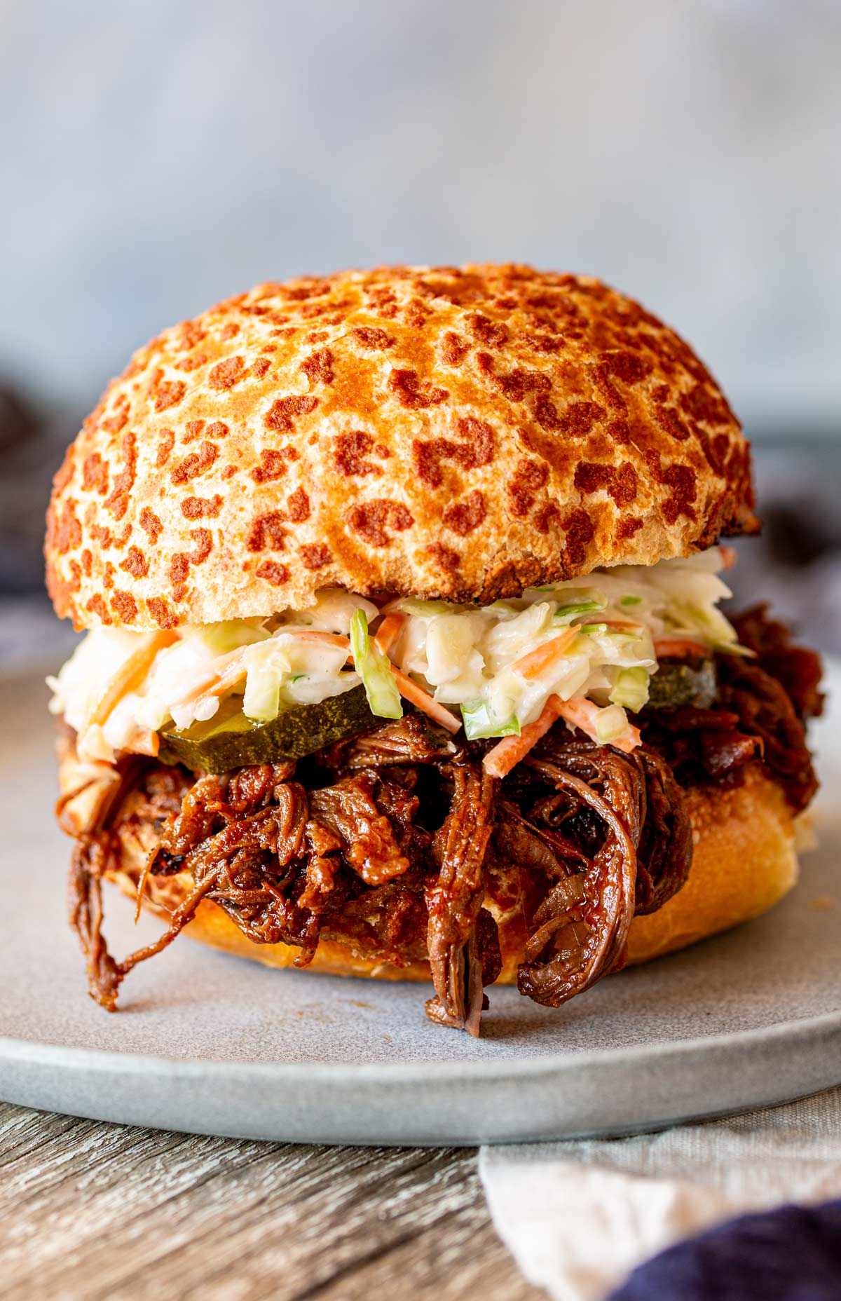 A BBQ beef sandwich featuring tender, shredded beef, sliced pickles, slaw, and a tiger bread roll, placed on a grey plate.