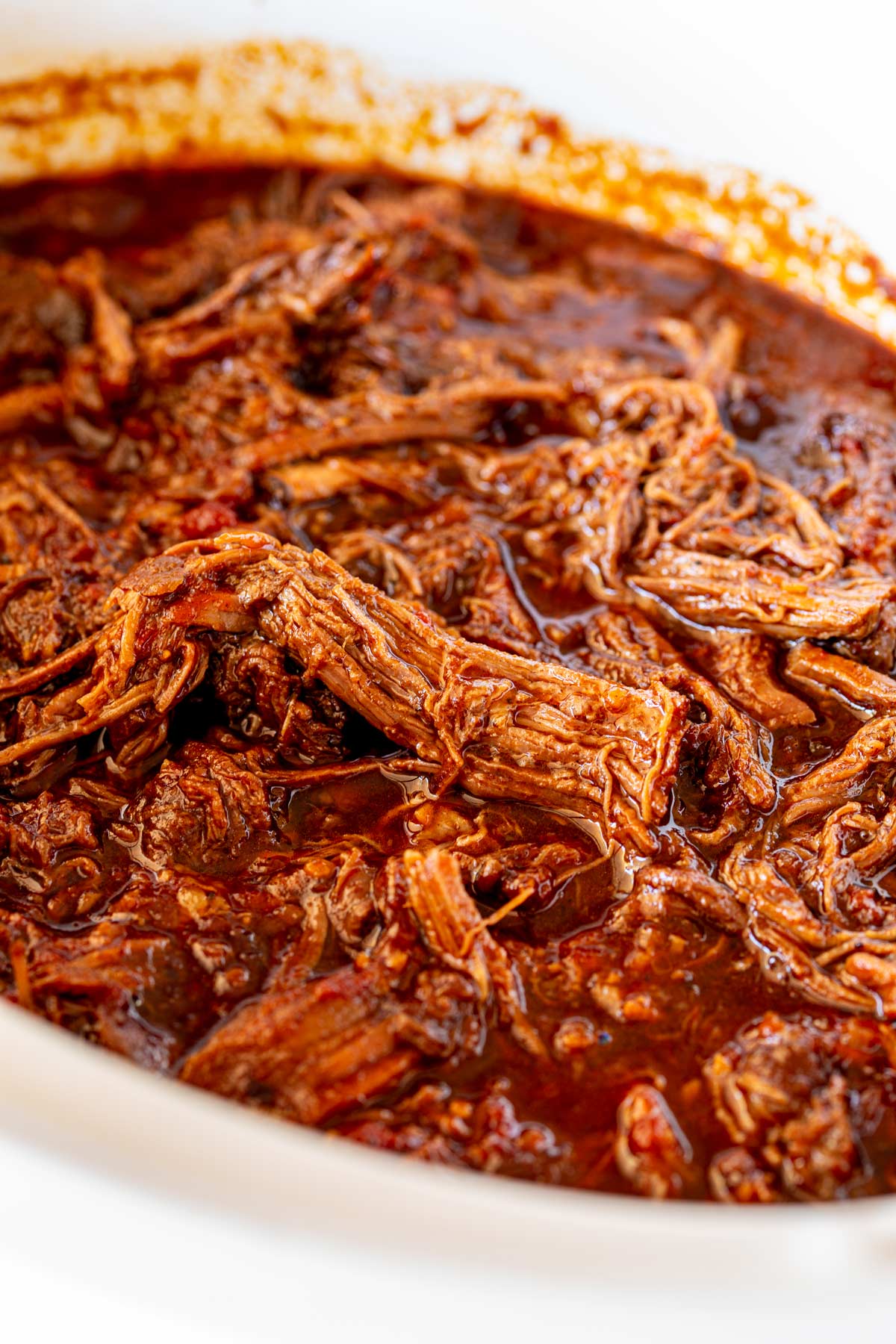 Shredded strands of bbef in a rich red BBQ sauce in the white bowl of a slow-cooker