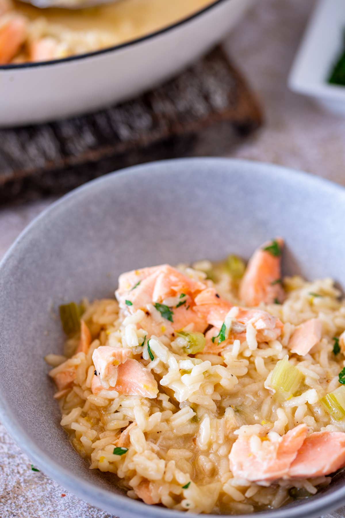 A grey bowl filled with risotto featuring small chunks of salmon, tender rice, and bits of green herbs, placed on a neutral-colored surface.