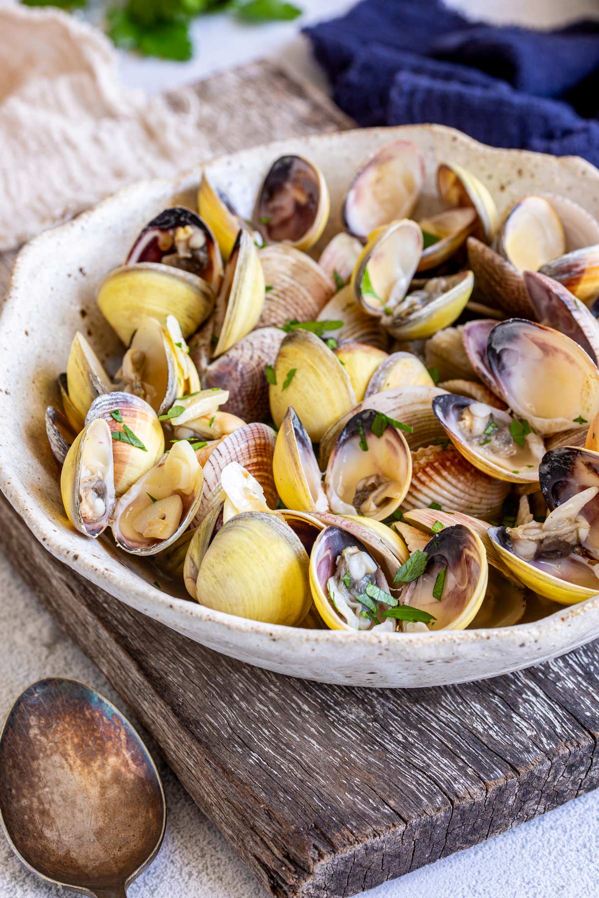 A stoneware bowl, sat on a rough wooden plank, holds a mixture of opened clams. The dish is topped with small pieces of parsley, with a rustic spoon in front and a blue napkin can be seen in the background.