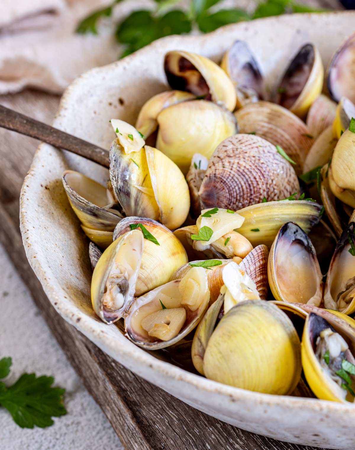 A close-up shot of cooked clams resting in a white bowl with a spoon in the bowl. The shells are golden and ivory colors, with a pop of green from the parsley sprinkled on top.