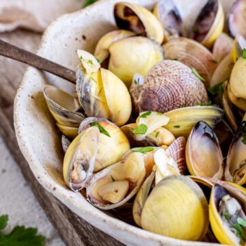 A spoon in a rustic bowl of freshly cooked clams garnished with parsley and garlic slices.