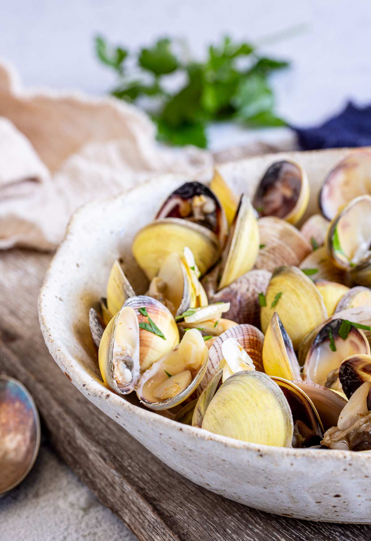 A white ceramic bowl filled with clams, their opened shells revealing the soft clam meat inside. The dish is garnished with chopped parsley, and the edges of a wooden board and a linen napkin are visible nearby.