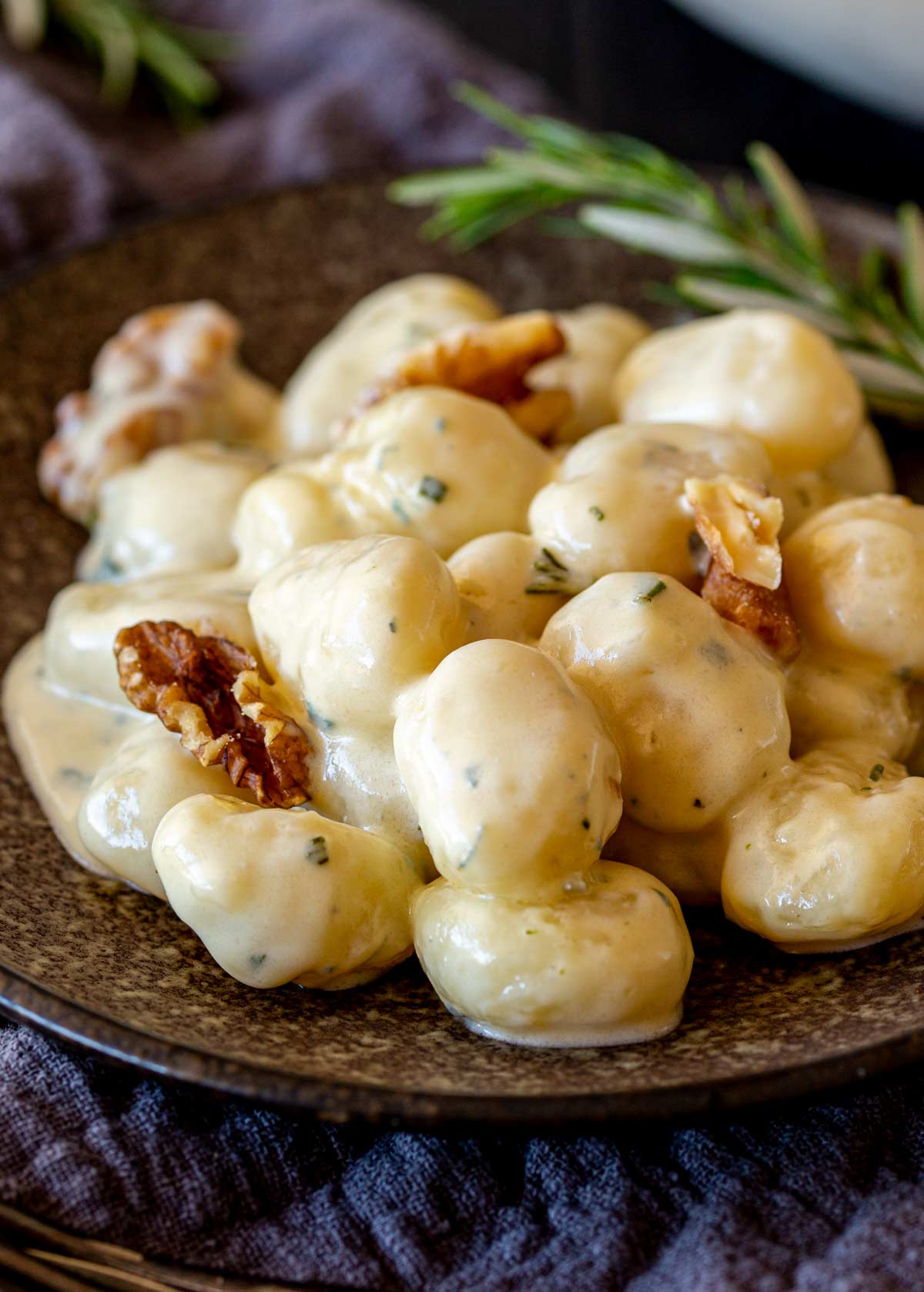 A bowl filled with gnocchi in a rich cream sauce, garnished with walnuts. A sprig of rosemary rests on the edge of the bowl, with a dark cloth napkin beneath.