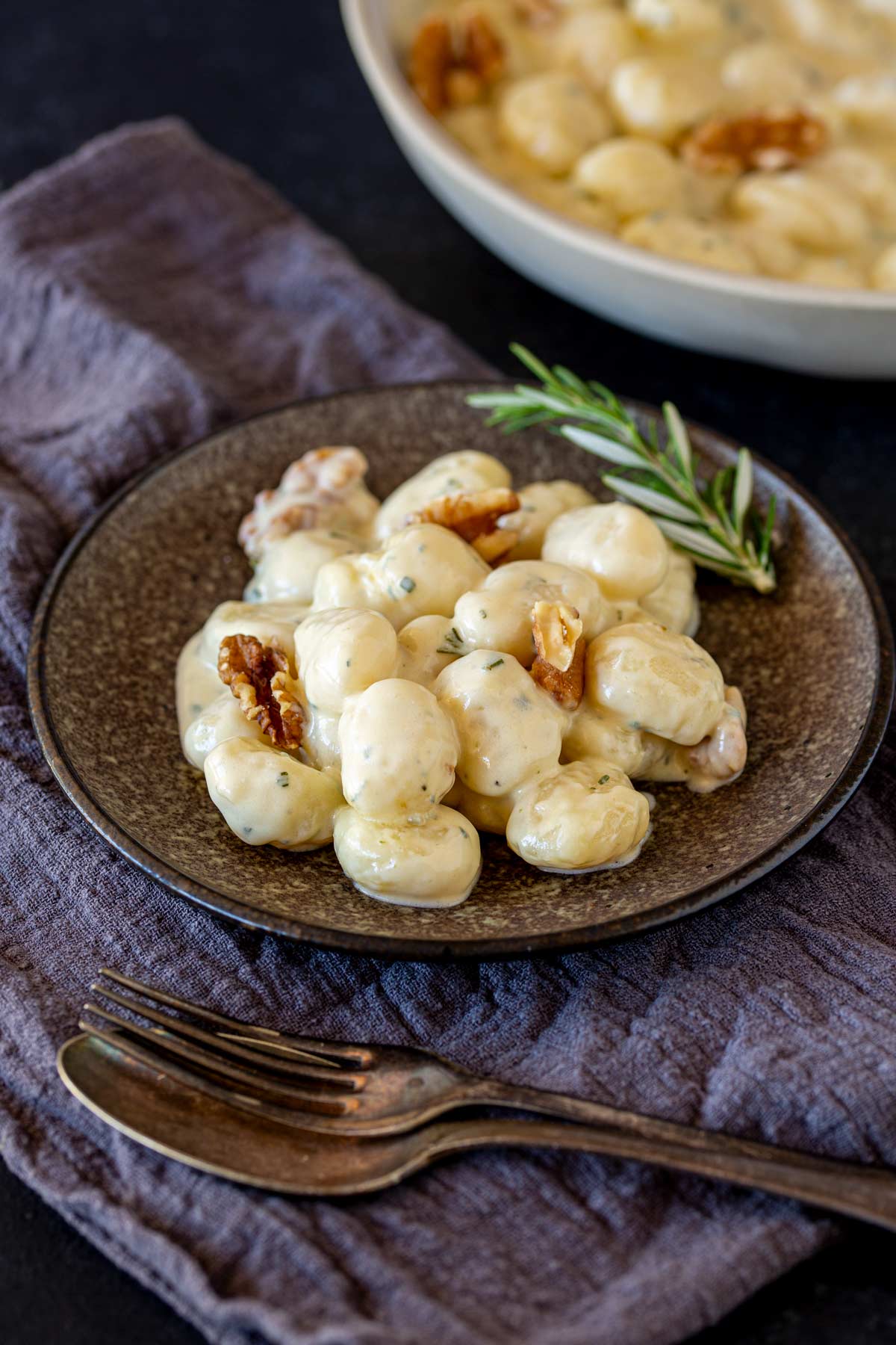 Gnocchi covered in creamy sauce, arranged on a brown plate, with walnut pieces for garnish. A rosemary sprig is placed beside the dish, and a dark cloth napkin sits underneath.