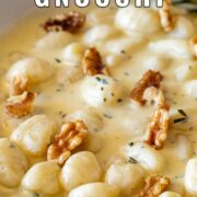 Pin Image: Gnocchi in a gorgonzola sauce with walnuts in a white skillet, with a rosemary sprig on the side. with text overlaid