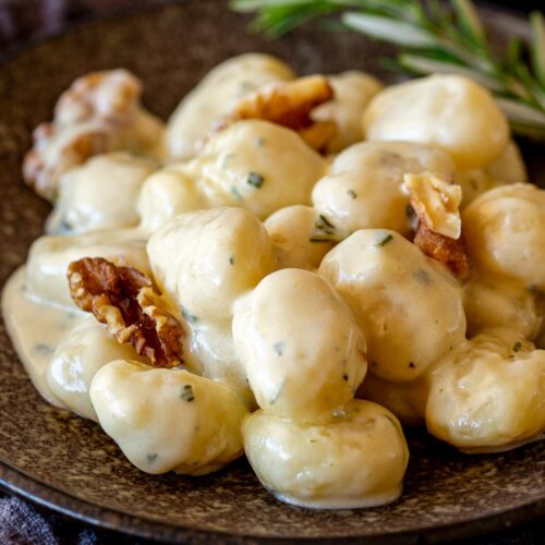 close-up of gnocchi covered in a creamy sauce, topped with walnuts, and served on a rustic brown plate. A sprig of fresh rosemary adds a touch of greenery in the background.