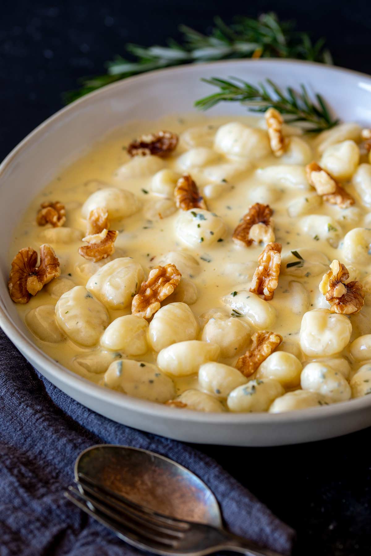 A white pan filled with gnocchi in a gorgonzola cheese sauce, with walnut halves added as garnish. A rosemary sprig rests on the side of the bowl, and a dark napkin lies beneath the setting.