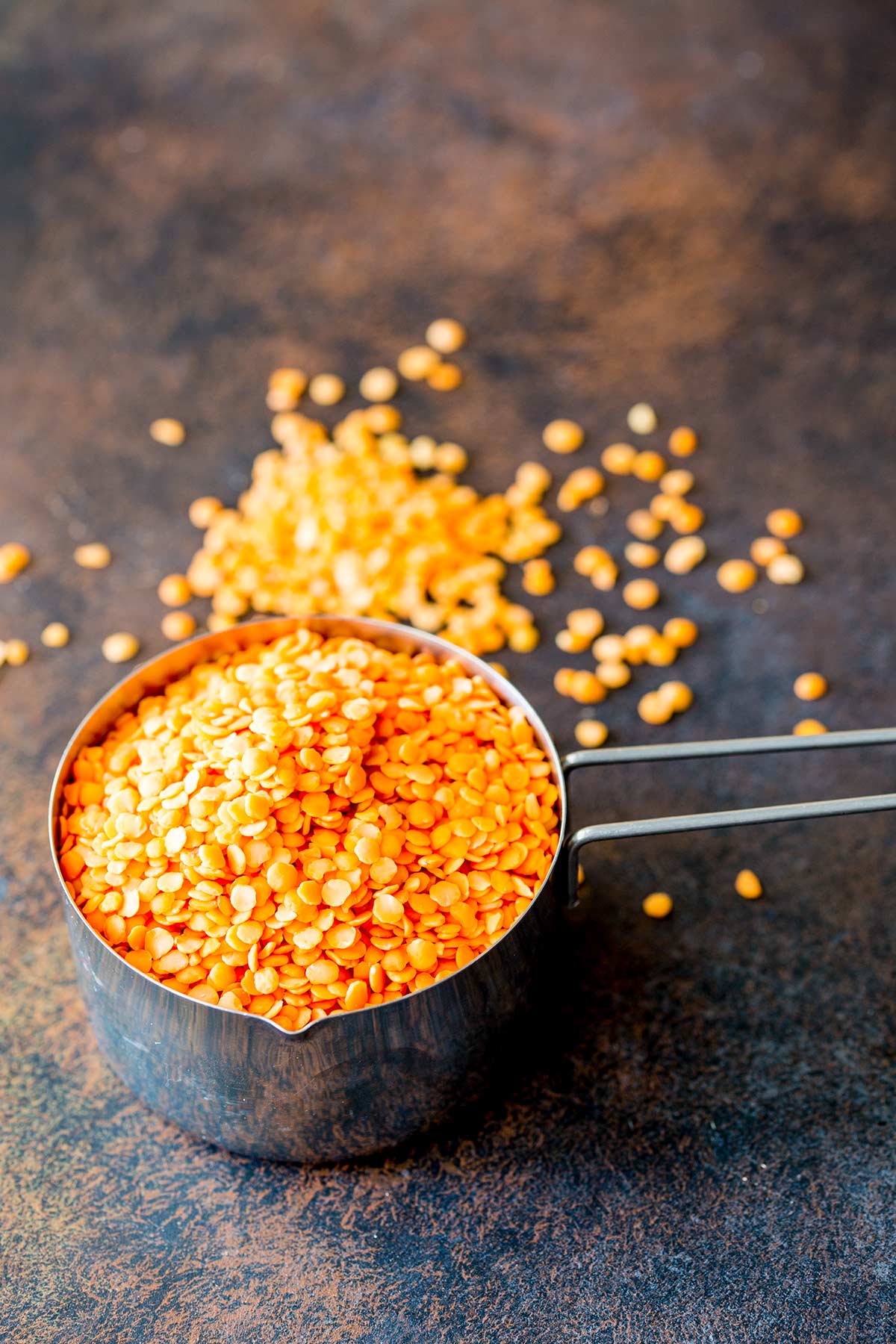 red lentils in a metal cup measure on a brown table with some lentils spilled out