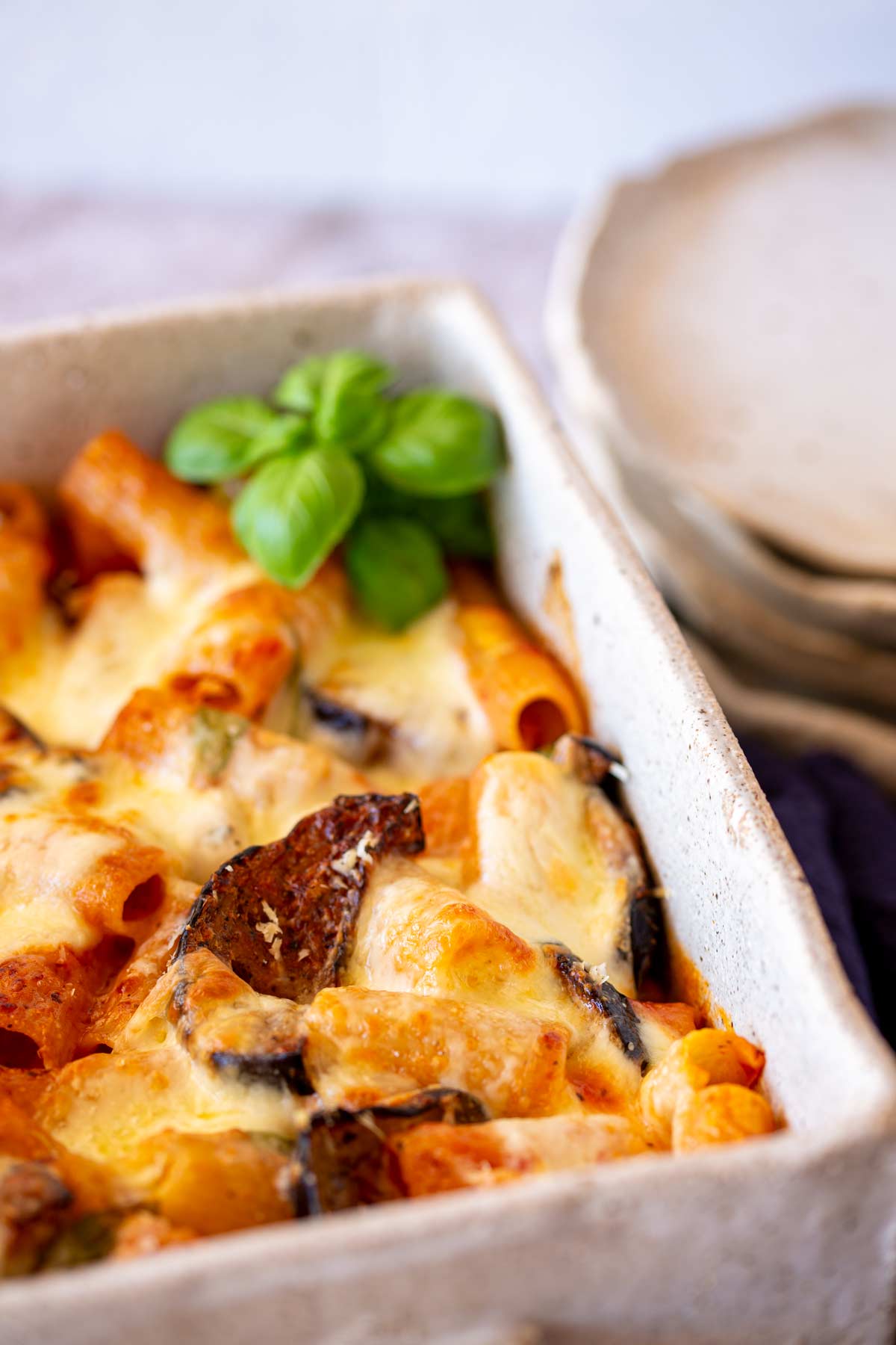 A close-up of baked pasta in a white casserole dish, with rigatoni, roasted eggplant, and golden-browned cheese, garnished with a sprig of fresh basil.