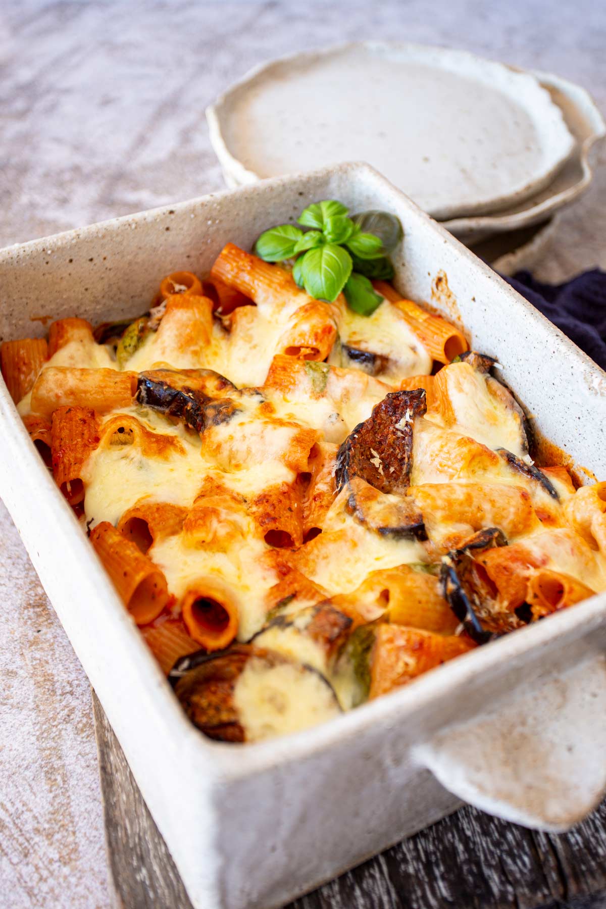 A ceramic baking dish on a rustic surface, showing baked rigatoni layered with slices of eggplant, tomato sauce, and melted cheese. Plates are stacked in the background.