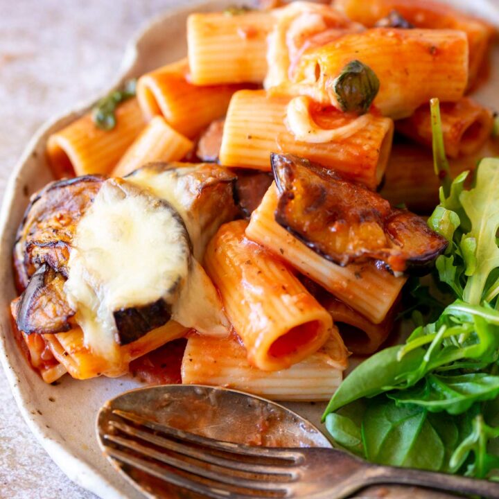 pasta tubes coated in tomato sauce, slices of roasted eggplant, and a sprinkle of melted cheese. There’s a small salad on the side, and a vintage-style fork rests nearby.