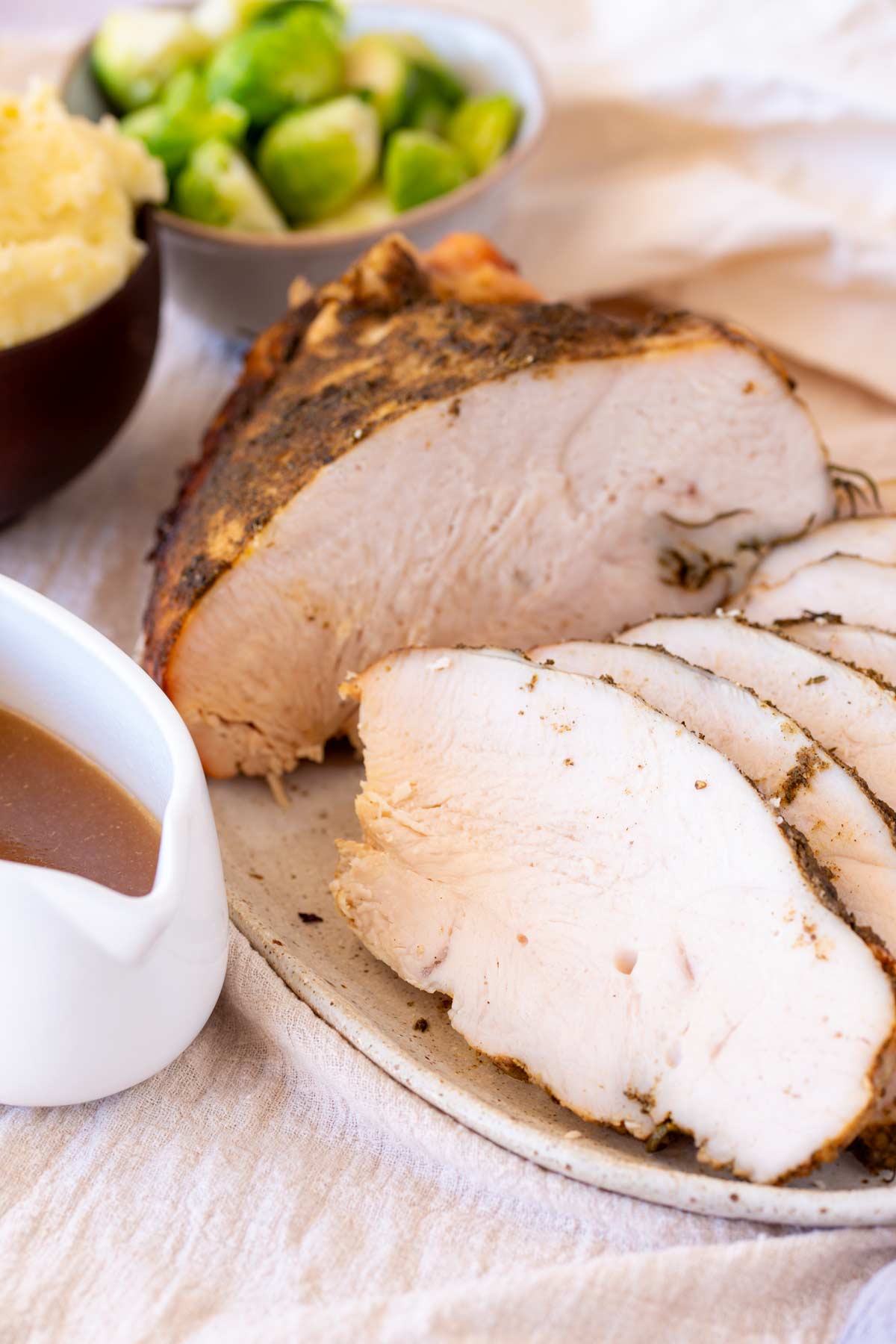 A serving plate showcasing slices of turkey breast, with a gravy boat next to it and vegetable behind