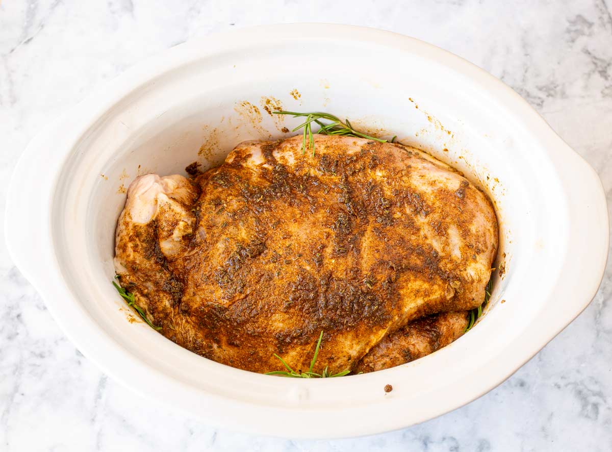 Raw turkey breast placed in a slow cooker with herbs and a sprinkle of seasoning on top.