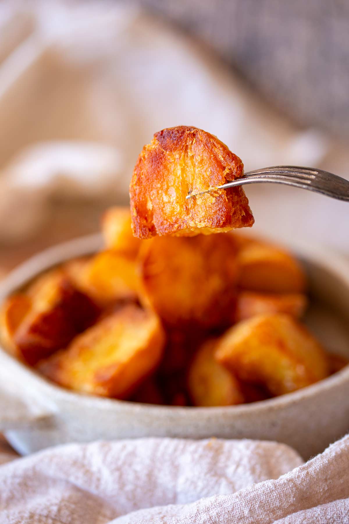 A roast potato held by a fork in focus, with a blurry bowl of crispy roast potatoes and a cream-colored napkin in the background.