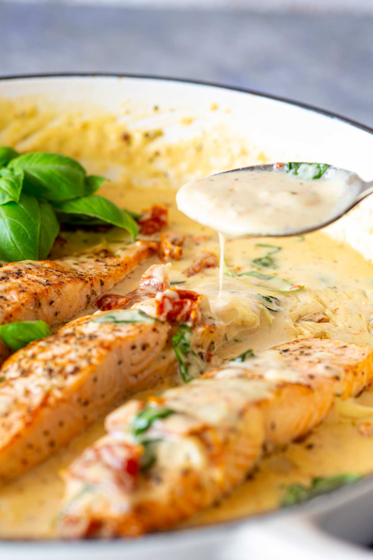 Close-up of a spoon pouring rich, creamy sauce over salmon fillets in a skillet. The creamy sauce is flecked with herbs and sun-dried tomatoes, with fresh basil adding a green garnish in the background.