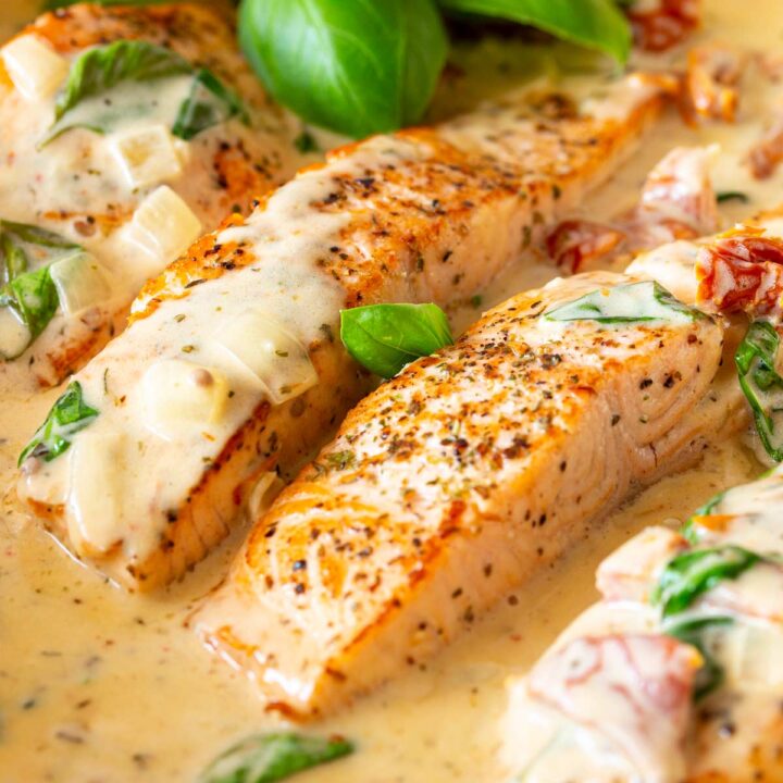 Close-up of salmon fillets cooked in a creamy sauce, garnished with fresh basil leaves and sundried tomatoes.