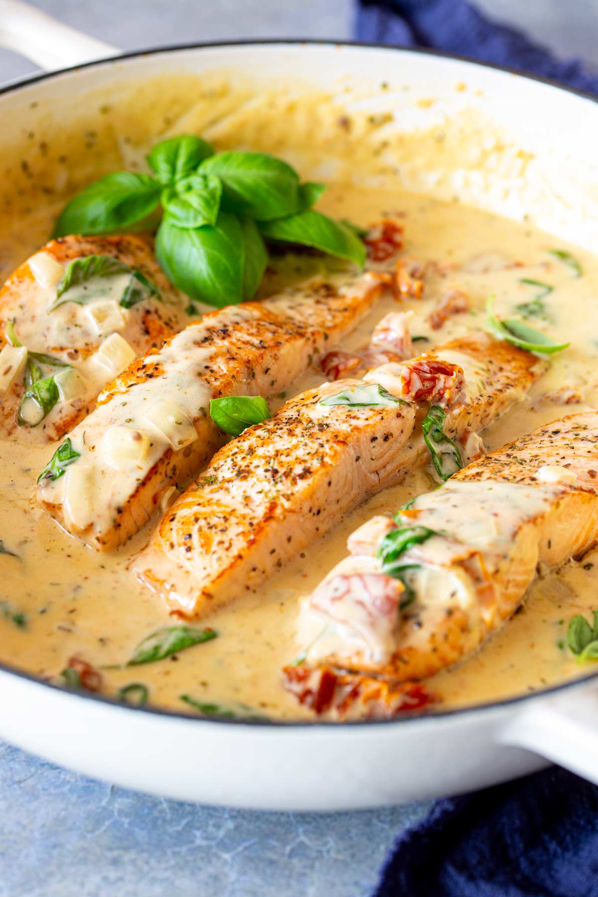 Salmon fillets nestled in a white skillet, covered in a creamy sauce mixed with basil and sundried tomato pieces. Fresh basil leaves add a finishing touch in the corner of the pan.