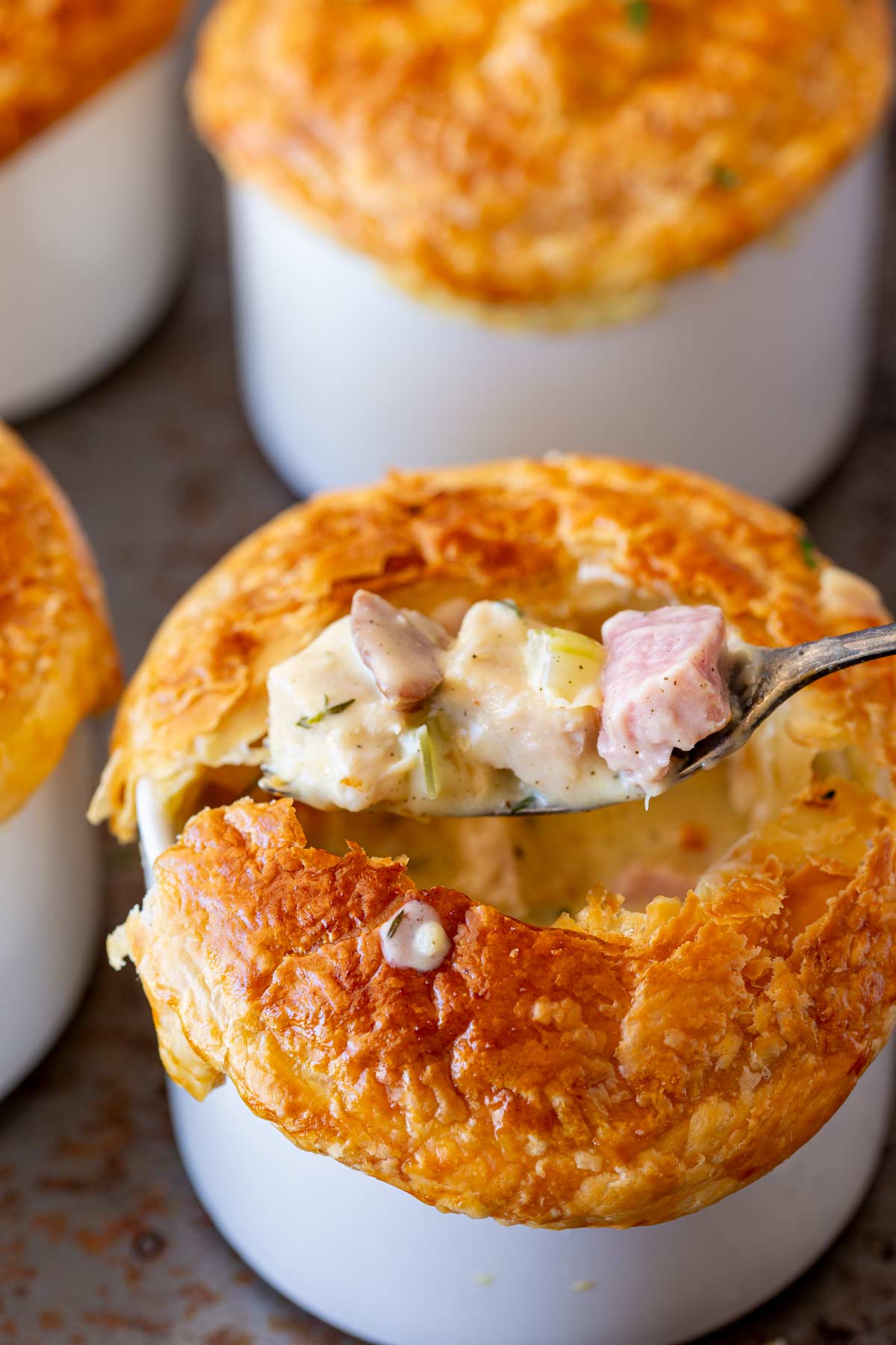 A spoonful of creamy turkey and ham pot pie filling is lifted from a white ramekin with a golden, flaky crust on top. In the background, additional ramekins are partially visible, showcasing their appetizing golden-brown tops.