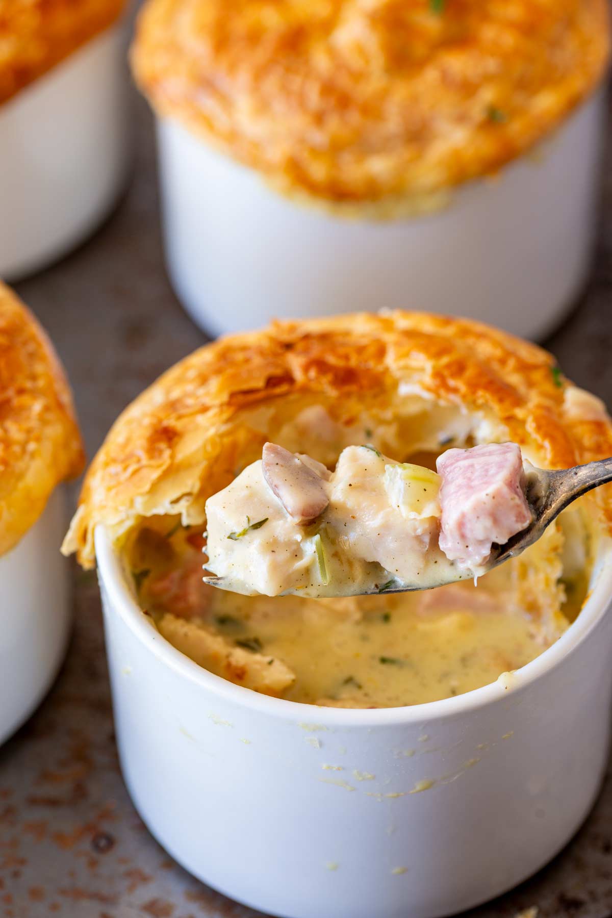 A spoonful of creamy turkey and ham pot pie filling is scooped from a white ramekin topped with a golden, flaky crust. More pies are out of focus behind.