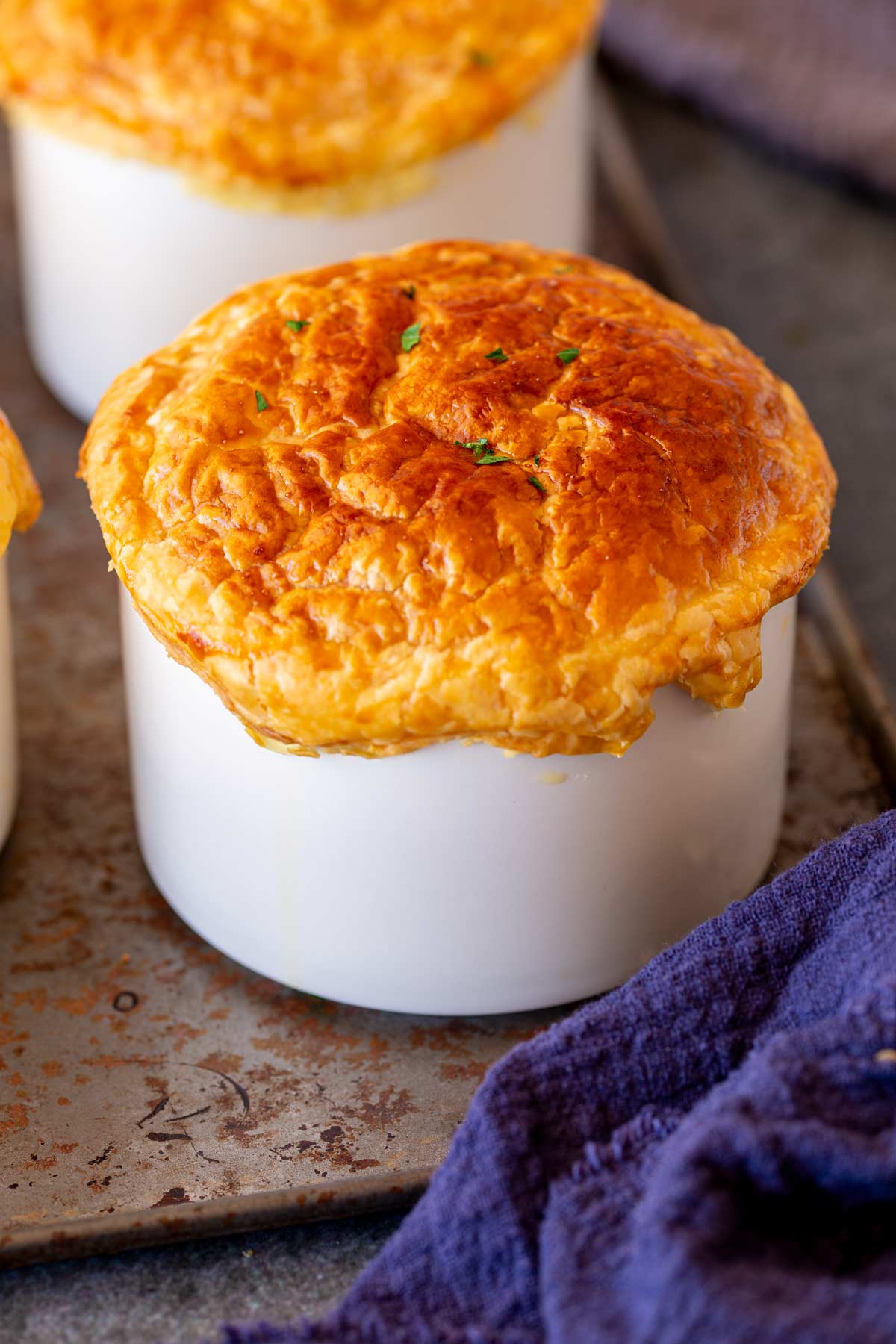 A golden-brown pot pie rises above the rim of a white ramekin, showcasing a flaky, puffed top with a few green herbs sprinkled on it. The ramekin sits on a rustic baking sheet, with a dark purple cloth partially visible in the foreground.