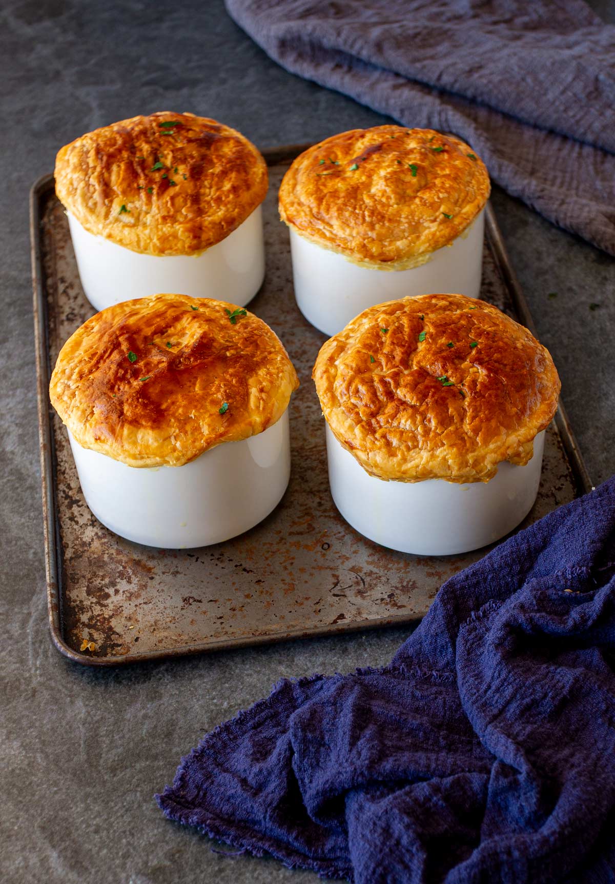 Four white ramekins with golden-brown baked pot pies on a metal baking sheet, placed on a grey surface with a dark blue cloth nearby.