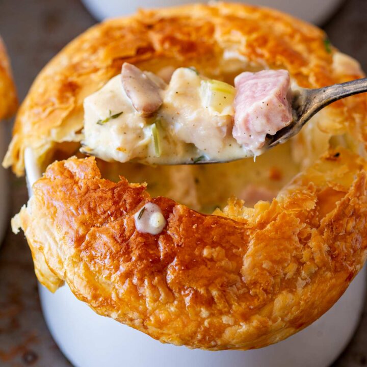 A spoonful of creamy turkey and ham filling is lifted from a white ramekin topped with golden-brown puff pastry.