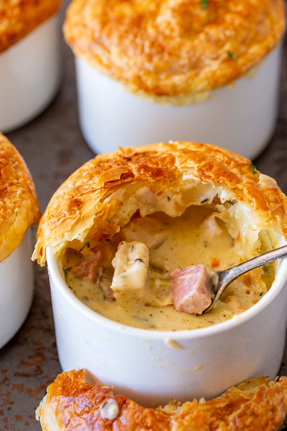 A close-up image of a pot pie served in a white ramekin featuring a golden, flaky pastry crust that is slightly cracked open at the top. Inside the ramekin is a filling with chunks of turkey and ham.