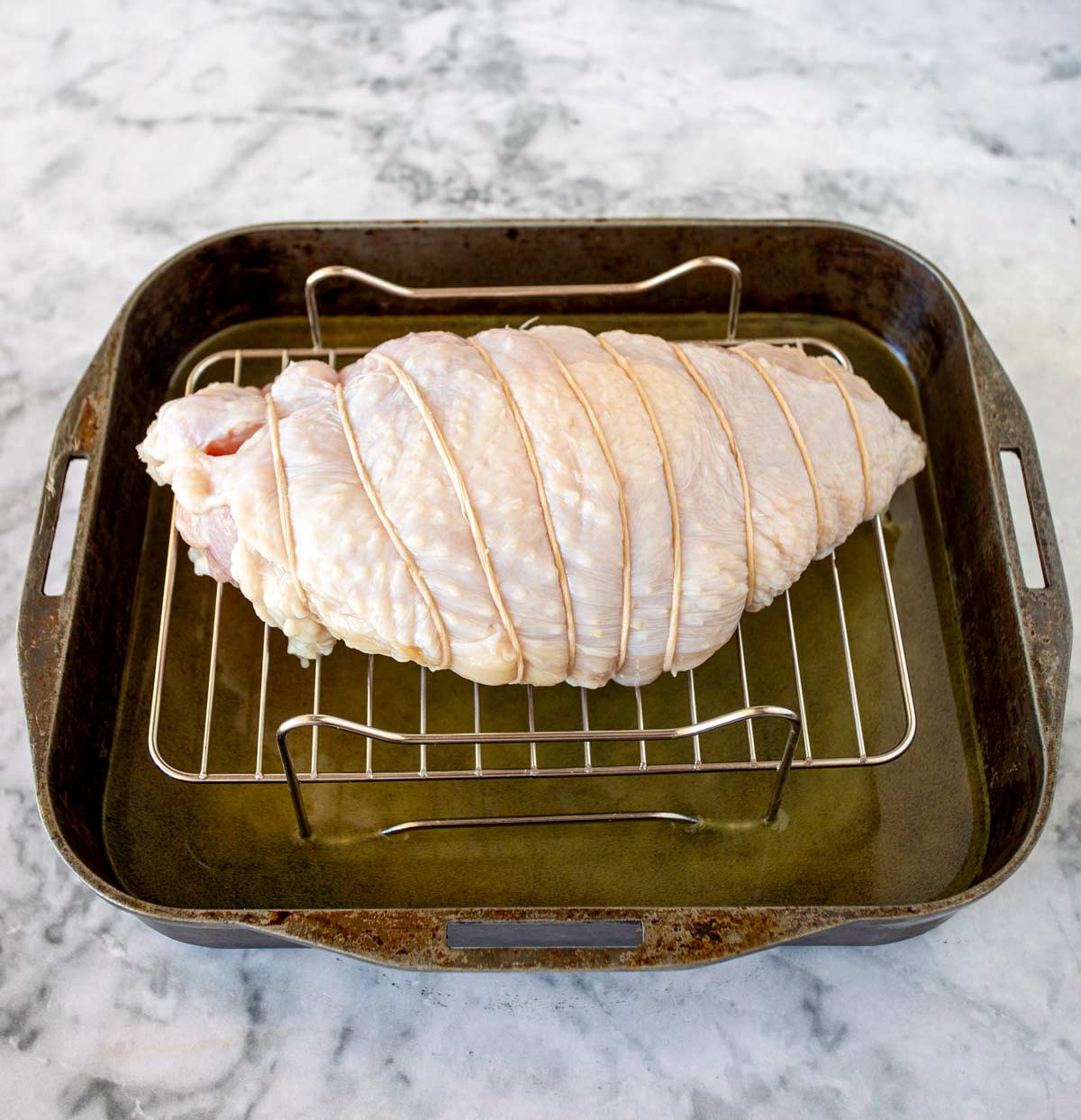 Boneless turkey breast tied with twine and positioned on a rack in a roasting pan, set against a marble countertop, ready for the oven.