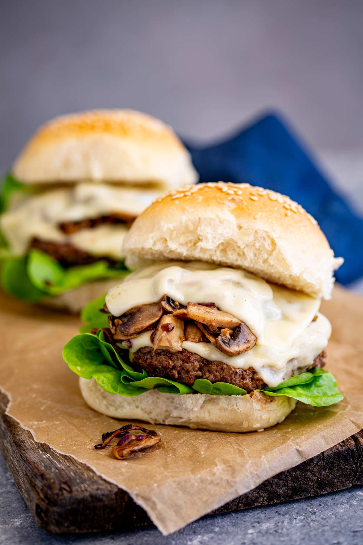 two burgers with a juicy patty, Swiss cheese, and sautéed mushrooms, set on a wooden board with a sesame seed bun slightly lifted to reveal the toppings.