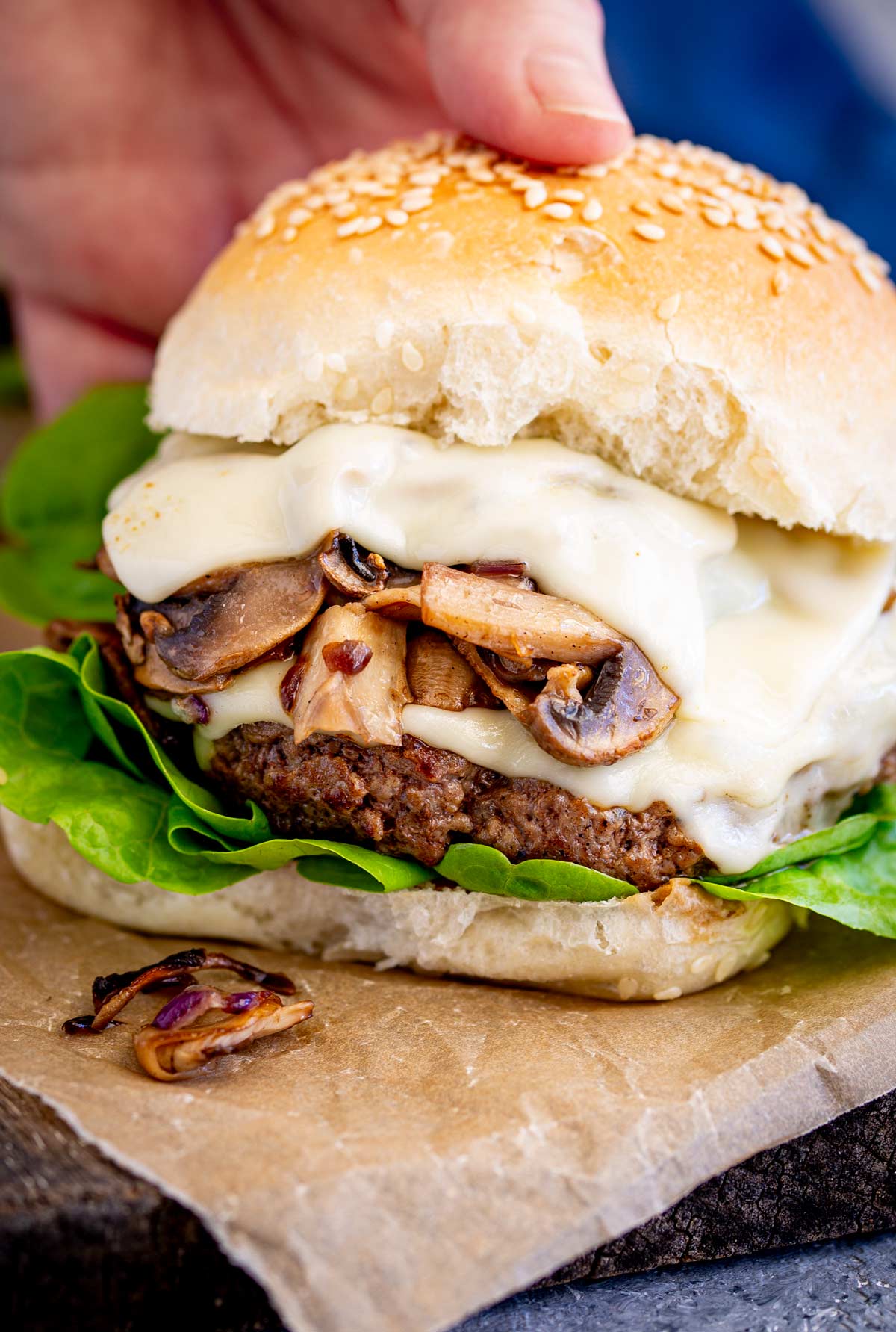 A hand picking up a Beef burger with melted cheese and mushrooms, shown on a rustic wooden surface.