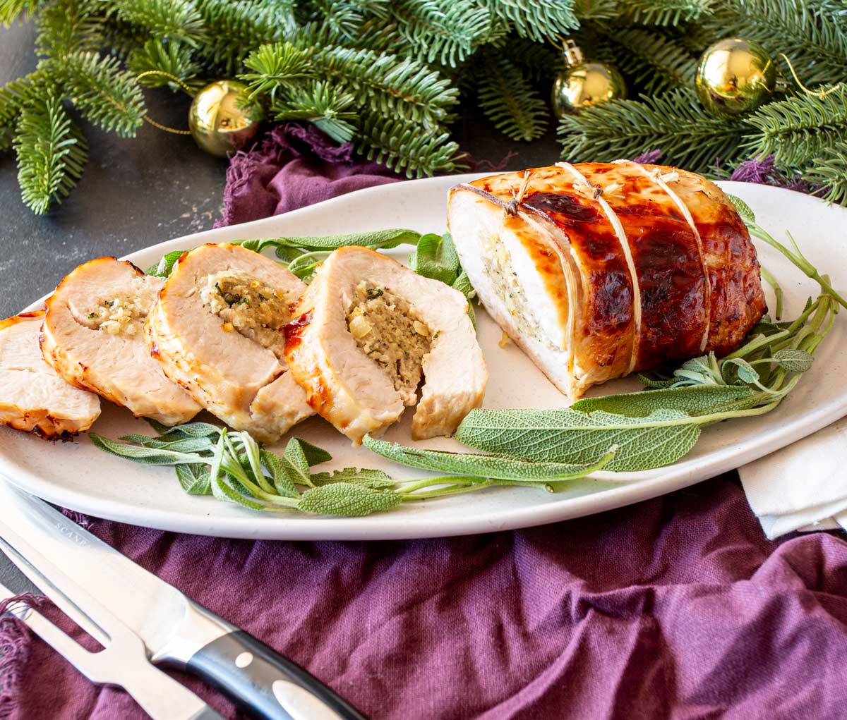 Rolled turkey breast with sage and onion stuffing, sliced and served on a white platter, garnished with fresh sage, surrounded by Christmas tree branches and gold ornaments.