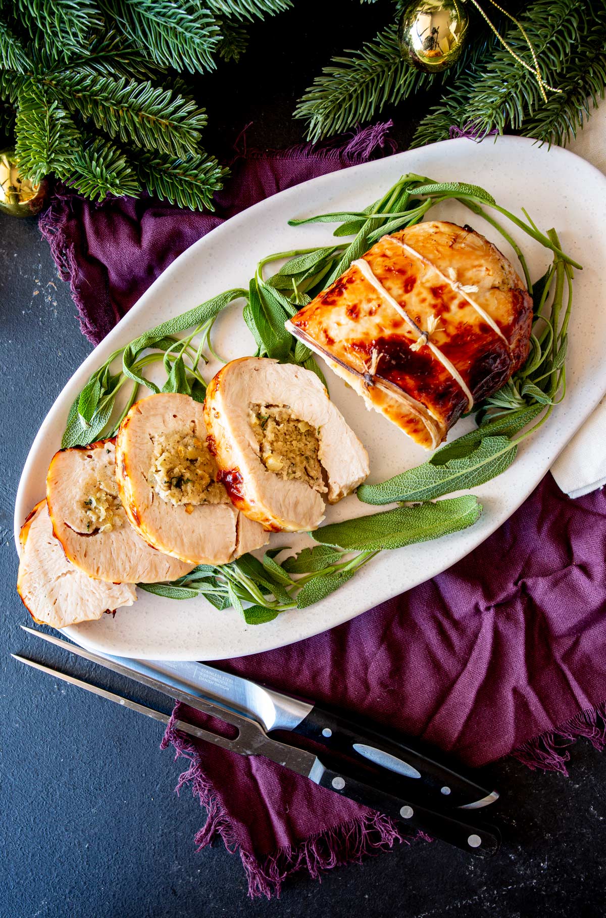 A top-down view of a rolled turkey breast stuffed with sage and onion, sliced and arranged on a white platter with fresh sage and a carving knife nearby.