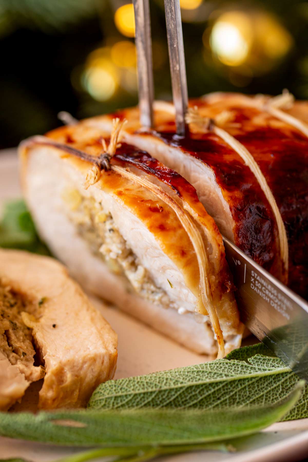 A knife cutting a slice of stuffed turkey breast, surrounded by fresh sage leaves and blurred golden holiday lights in the background.