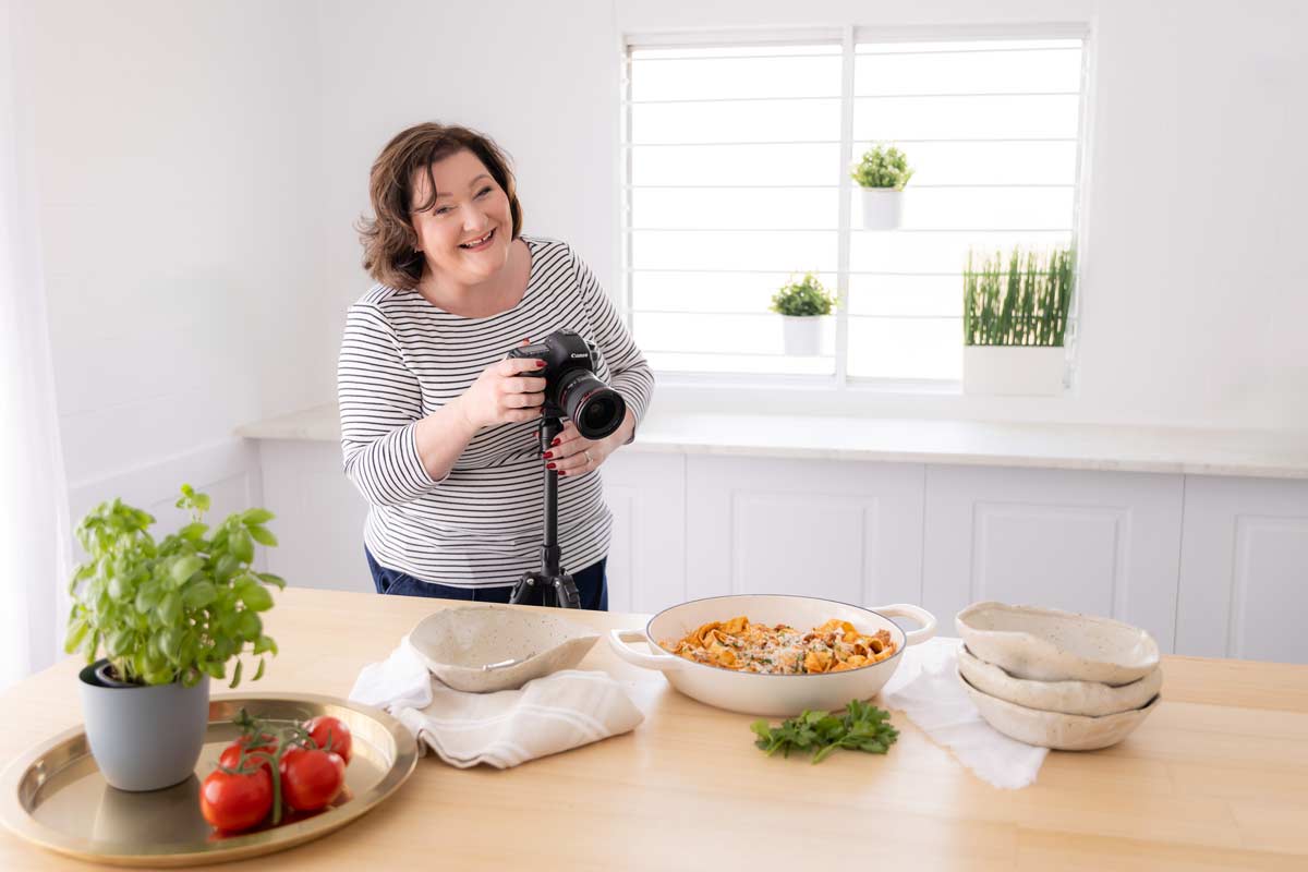 a lady laughing while pointing a camera on a tripod as a dish of food