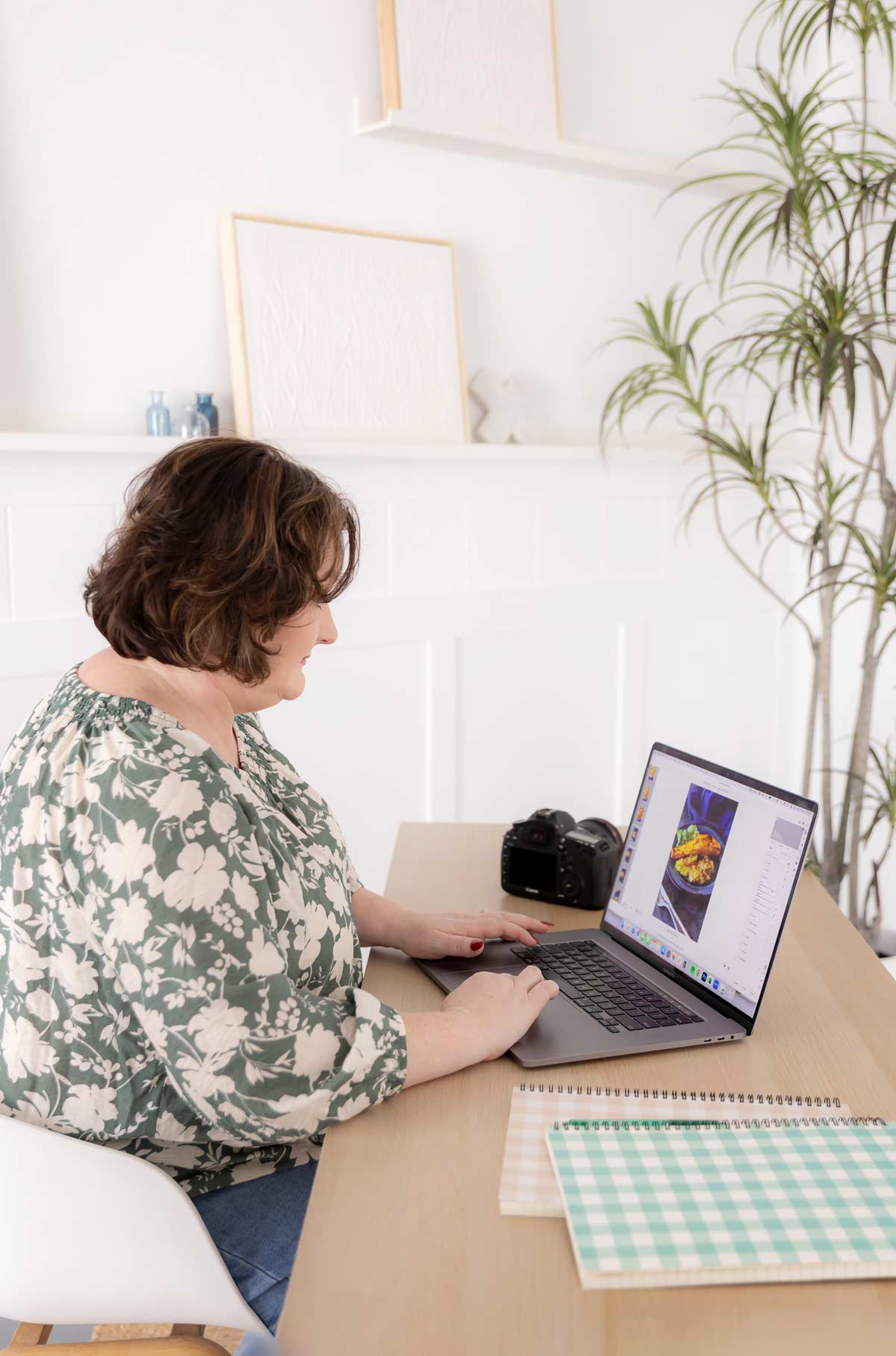 a lady in a flowery top sat at a desk with a laptop, exercise books and a camera