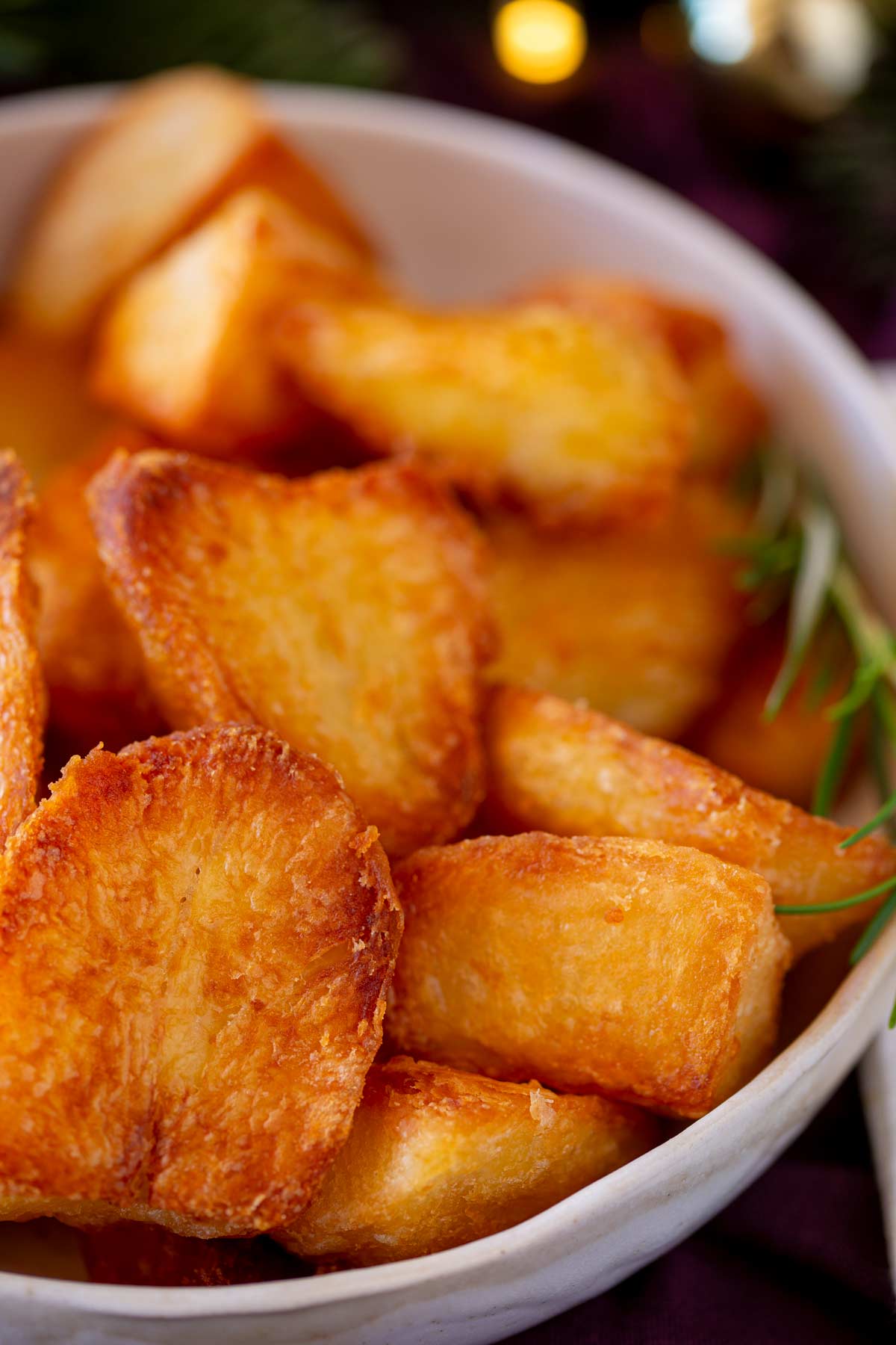 Golden roast potatoes in a white dish with a sprig of rosemary