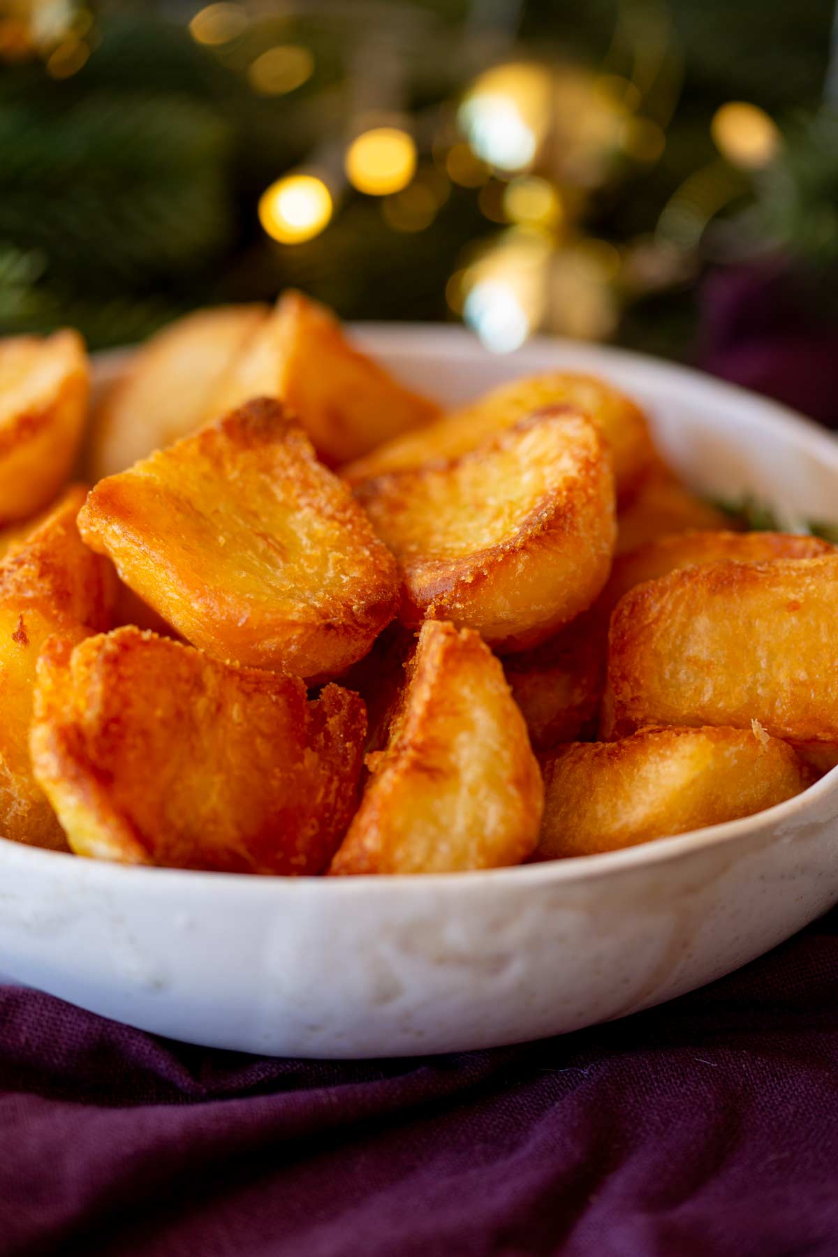 Golden, crispy roast potatoes served in a white dish, on a table with a purple napkin