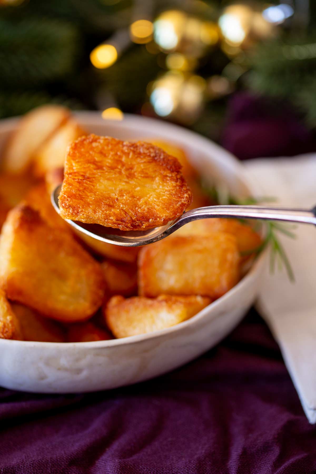 a spoon lifting a roast potato from a white bowl, with Christmas foliage blurred in the background