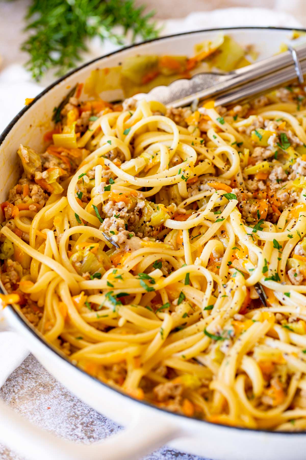 A white cast iron pan filled with pasta coated in a buttery ground pork sauce. The rustic surface and cream linens frame the dish, with rosemary sprigs in the background.