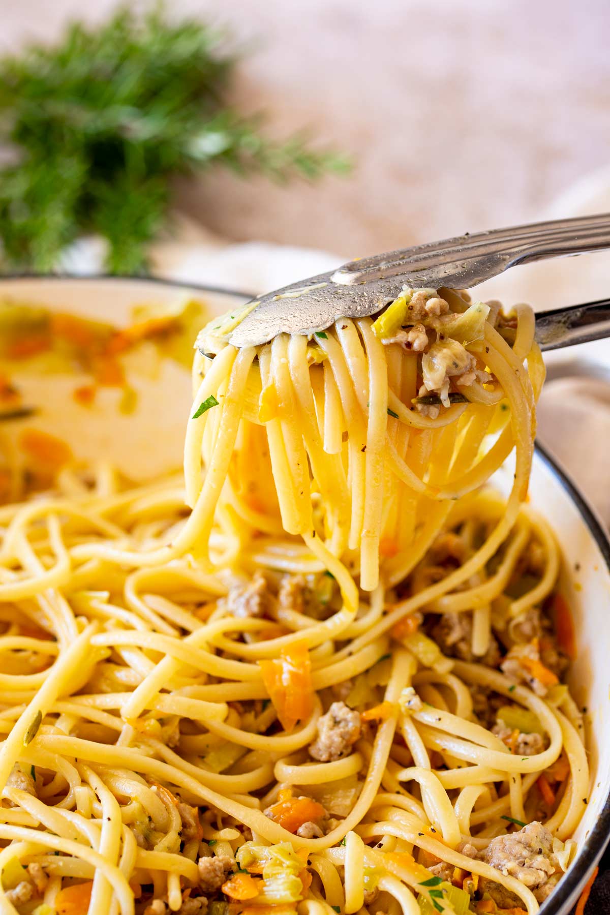 Tongs lifting a portion of pasta from a white cast iron pan, showcasing the buttery ground pork sauce clinging to the pasta.